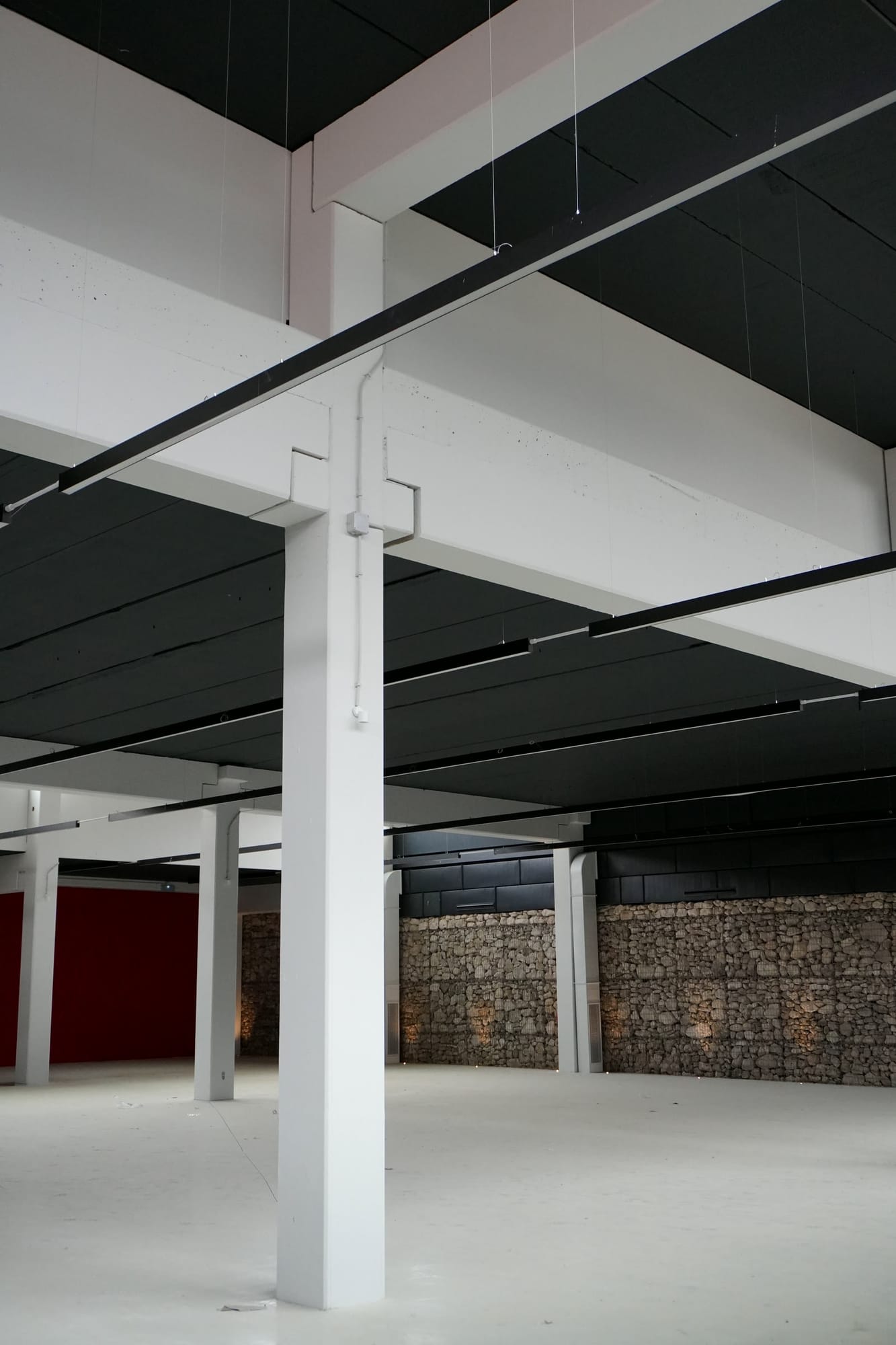 Nave diáfana con pilares blancos, techo oscuro con lámparas lineales y muro de gaviones de piedra