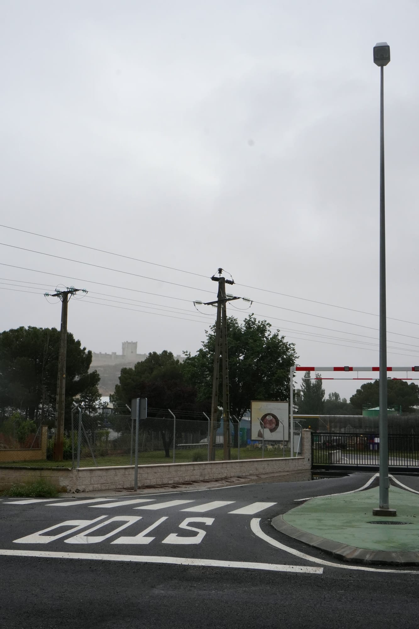 Entrada asfaltada con isleta, farola y marca de STOP, con la barrera de acceso a la fábrica al fondo