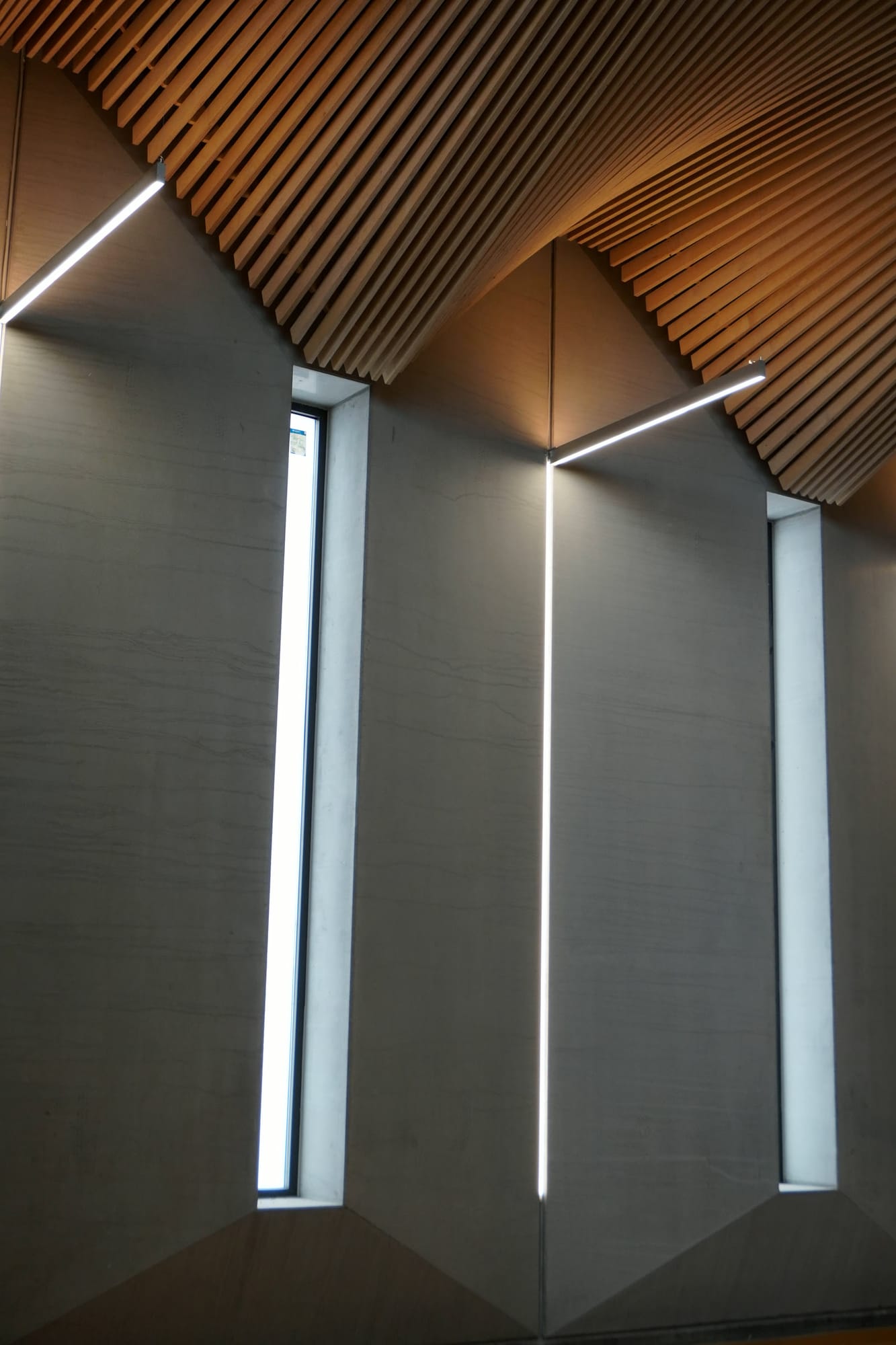 Muro gris con ventanas verticales y líneas de luz bajo un techo de lamas de madera