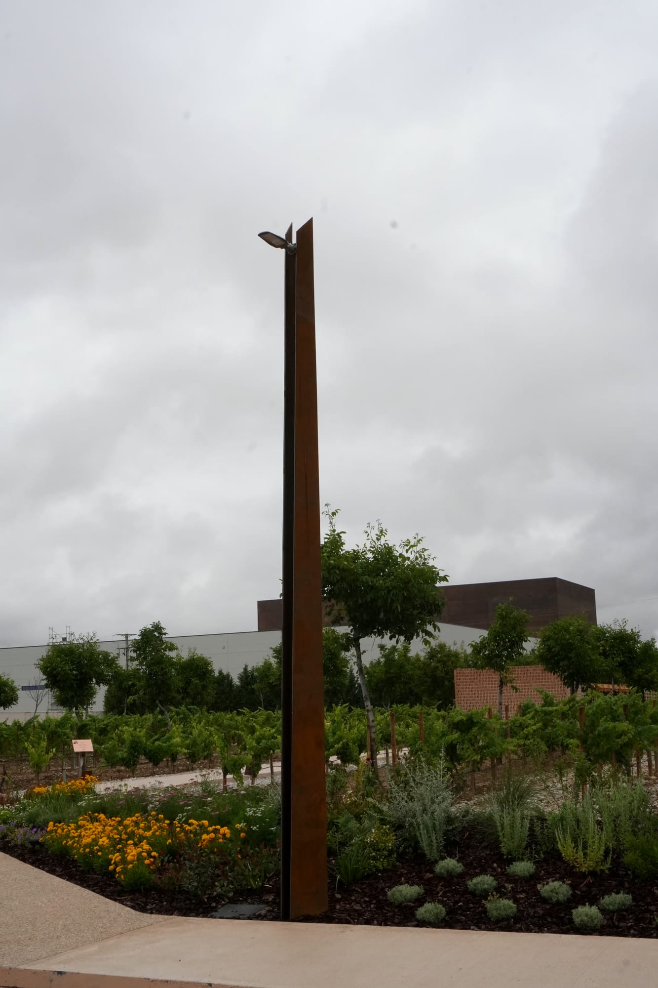 Poste alto de acero corten en el jardín de viñas, con la bodega al fondo